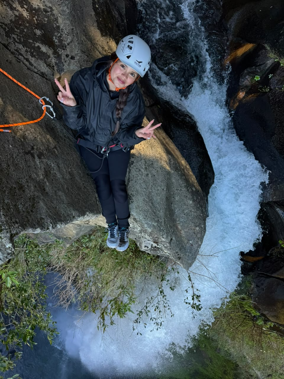 Participante con casco haciendo el signo de la paz sobre una roca junto a la cascada