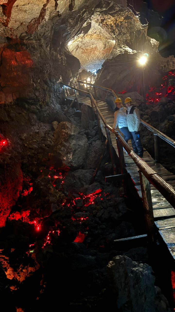 Dos visitantes con casco sobre la pasarela de madera de una cueva volcánica iluminada en rojo