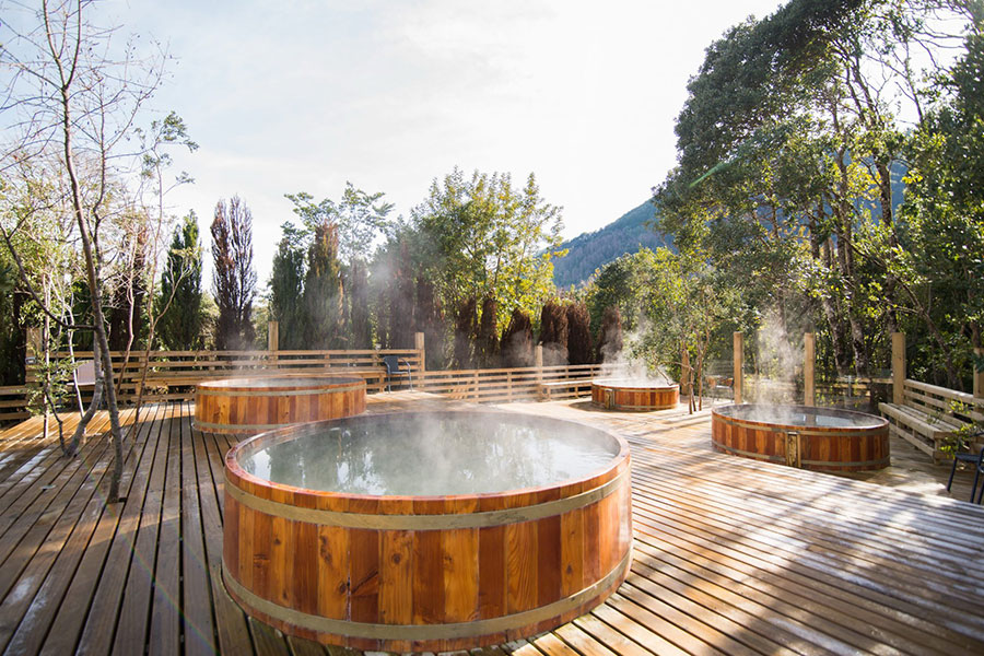 Tinajas de madera con aguas termales humeantes sobre una terraza rodeada de bosque y montaña